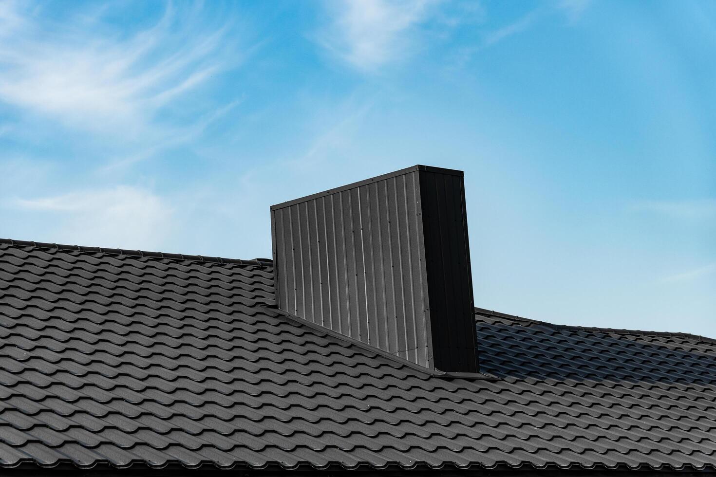 A modern, black chimney sits atop a black, tiled roof. The chimney is rectangular with vertical lines. The background is a blue sky with white clouds photo