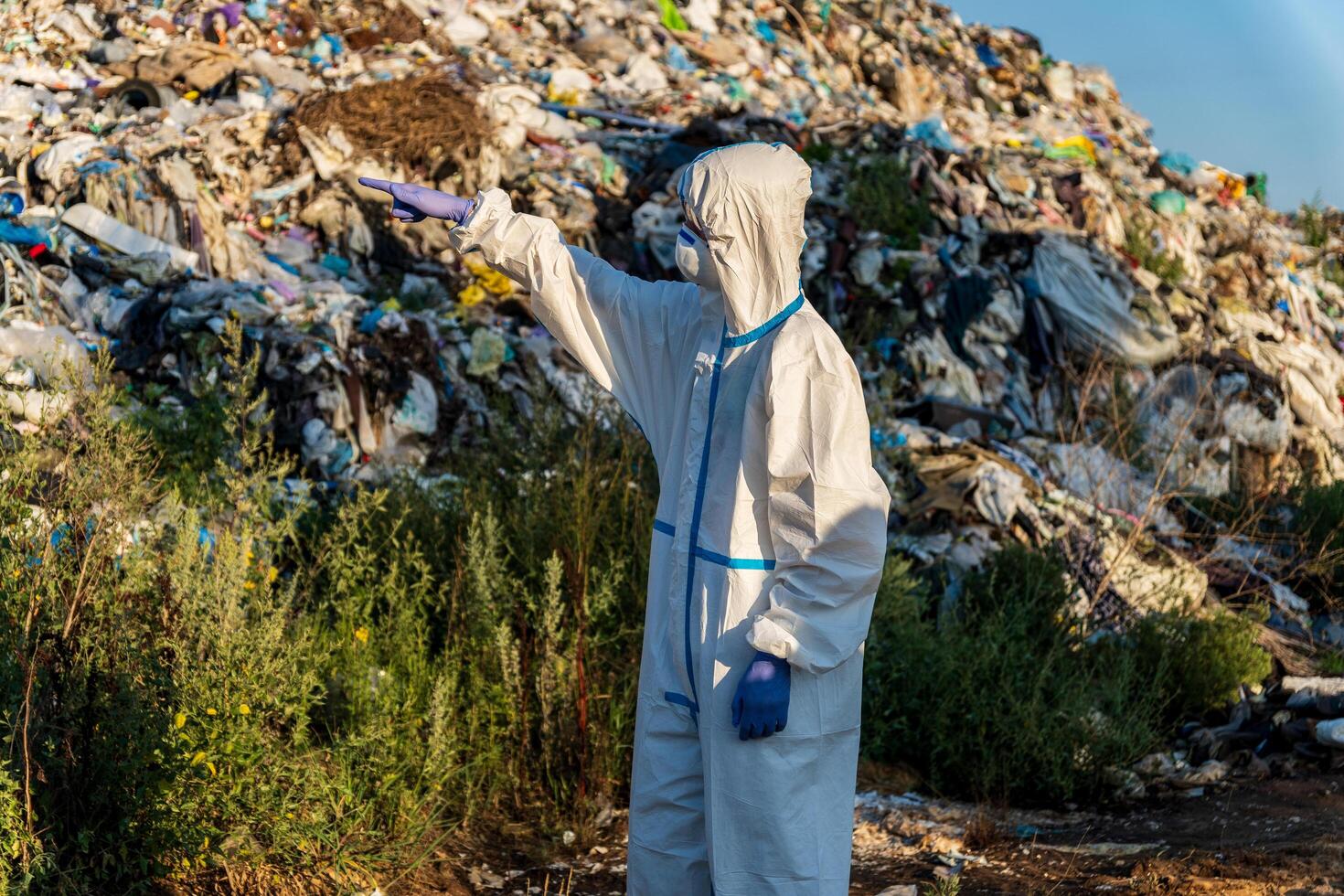An environmental worker dressed in protective gear surveys a landfill filled with waste while pointing at the surroundings under clear skies photo