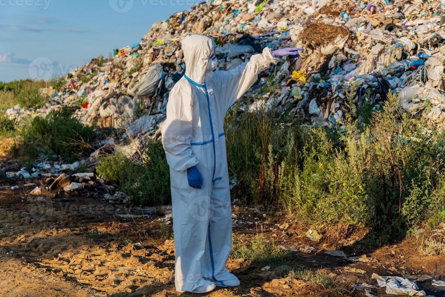 Wearing full protective clothing and gloves, a worker examines a large landfill, meticulously checking for any hazardous materials under the clear blue sky photo