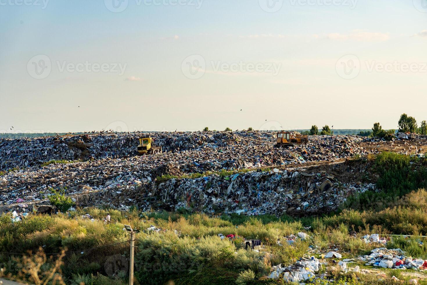 Bulldozers maneuver through a substantial landfill, compacting trash under a clear afternoon sky, surrounded by sparse vegetation and distant trees photo