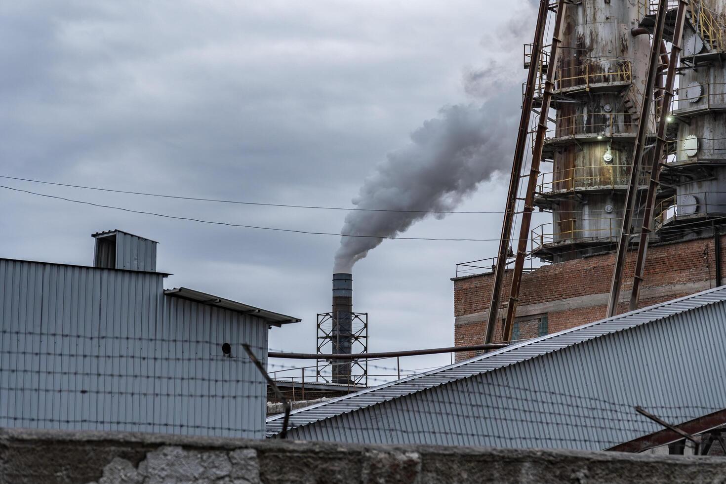 A factory stands prominently, releasing smoke into the overcast sky, surrounded by industrial structures and rooftops, showcasing an urban environment photo