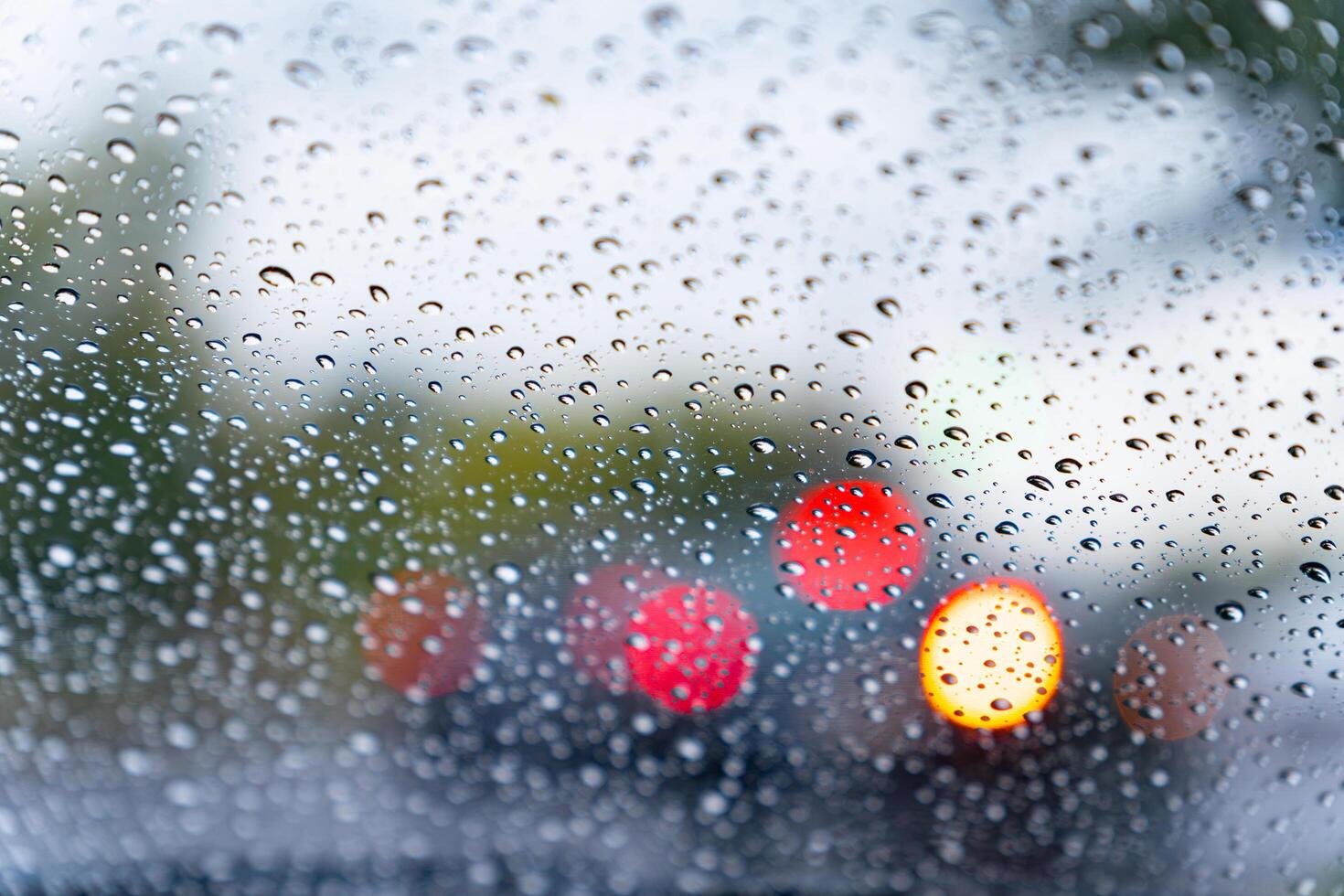 Raindrops cling to a car windshield, distorting the view of bright red and yellow lights from vehicles moving in the rainy city. The atmosphere feels moody and reflective photo
