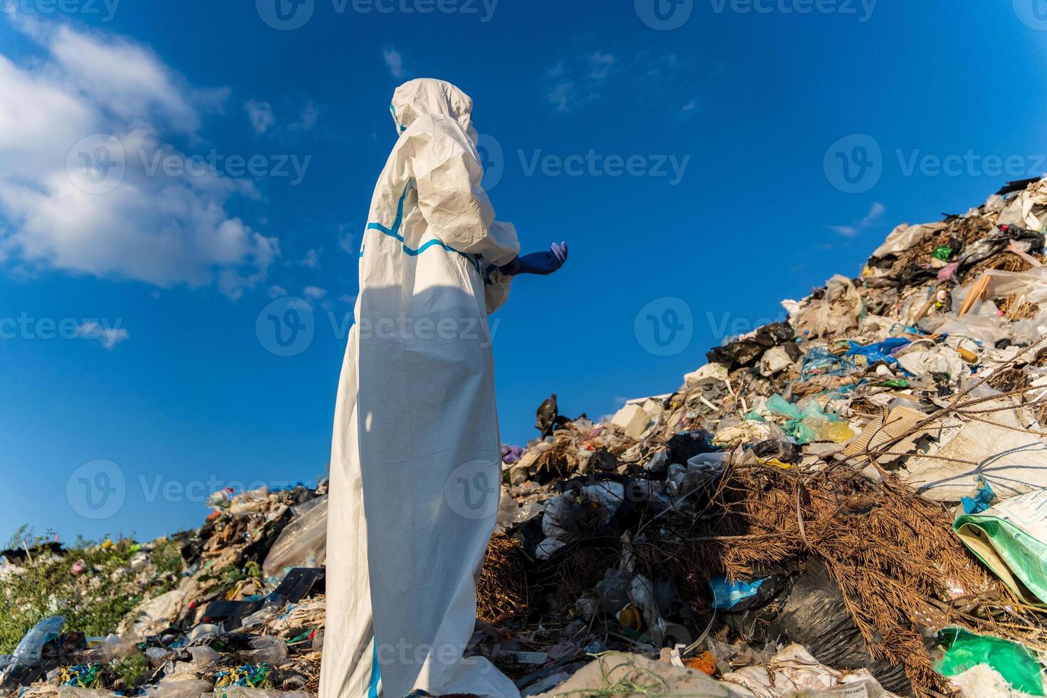 A waste management worker in protective clothing inspects a landfill filled with various types of debris under a sunny blue sky in the daytime photo