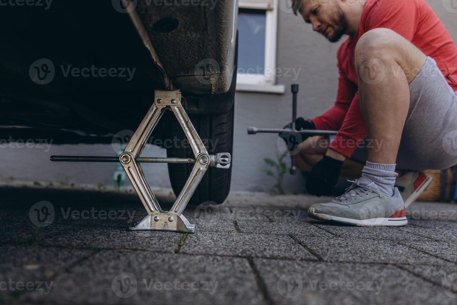 Close-up of man changing a car tire in front of his house. The vehicle is lifted with a jack, tools lie nearby. The scene captures a hands-on repair moment in daylight photo