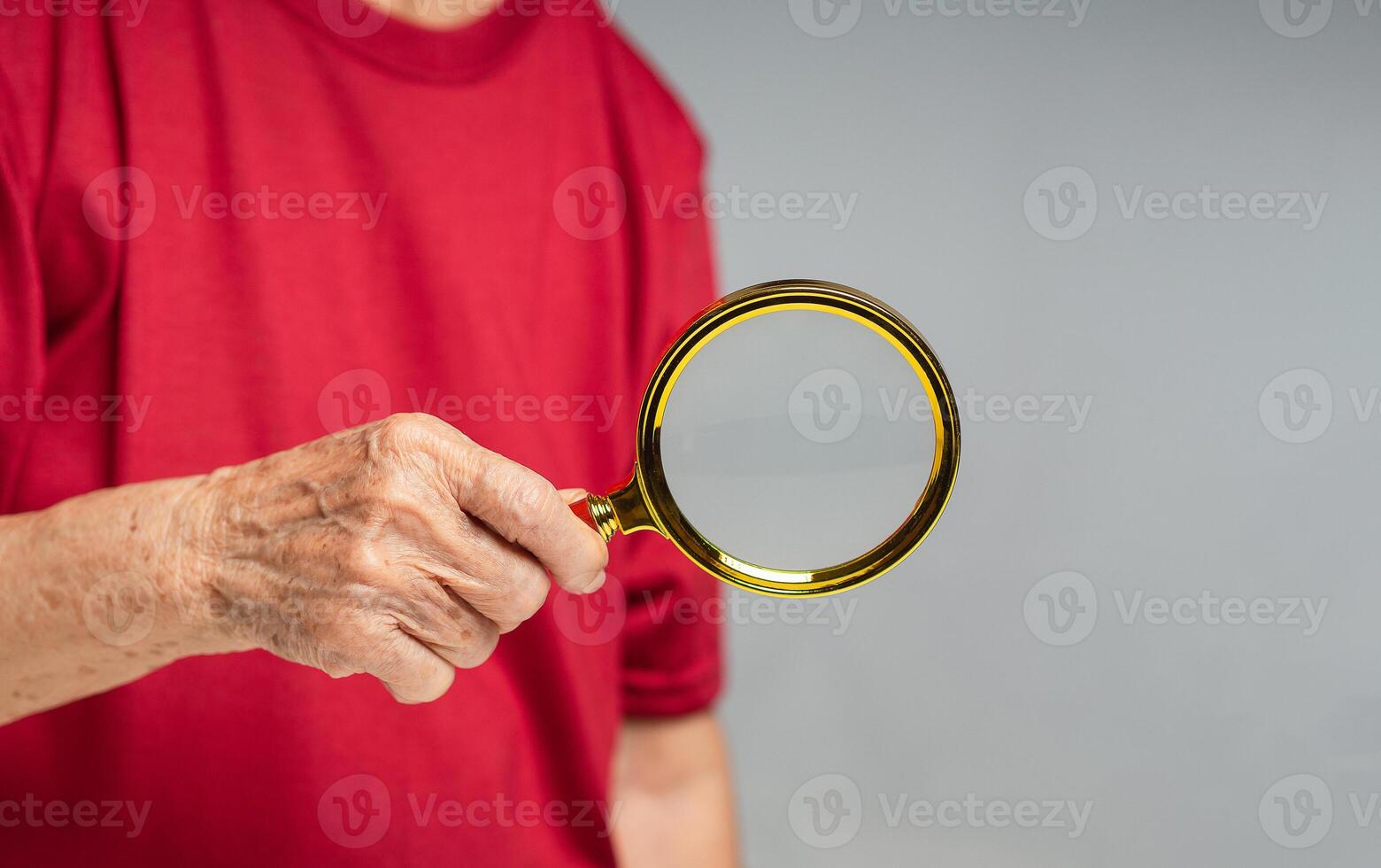 Close-up of a seenior woman wearing a red casual holding a magnifying glass while standing against a gray background. photo