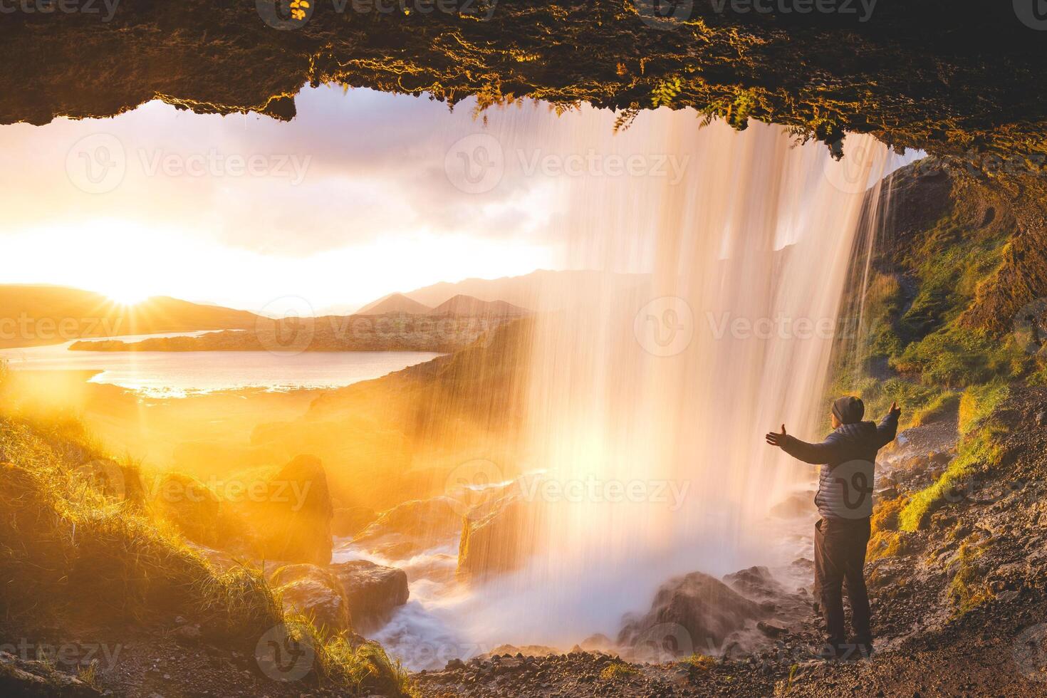 Man enjoys the view of a magical sunset from behind the Selvallafoss waterfall on the Snaefellsnes Peninsula in western Iceland, standing beneath the cold, clear water. Nature treasure photo