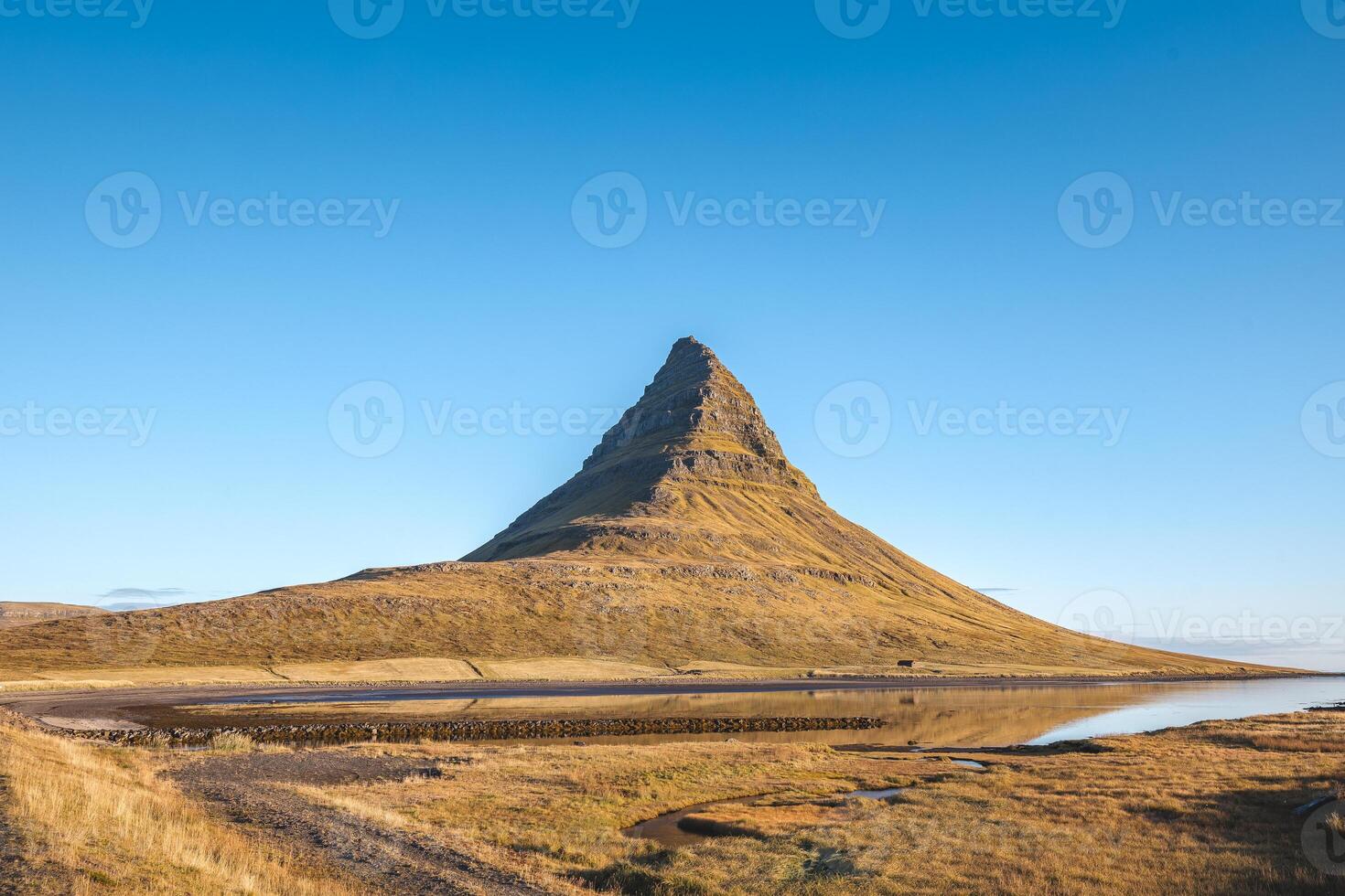Peak of Kirkjufell rises sharply on Snaefellsnes peninsula Iceland bathed in golden sunlight with a still lake and cloudless sky completing this perfect view photo