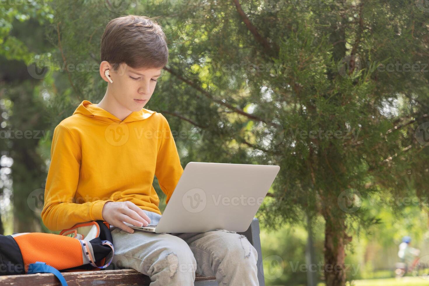 A teenage boy in a yellow hoodie sits in a park with a laptop, wearing wireless headphones. Green trees in the background create a natural summer setting. Side view. photo