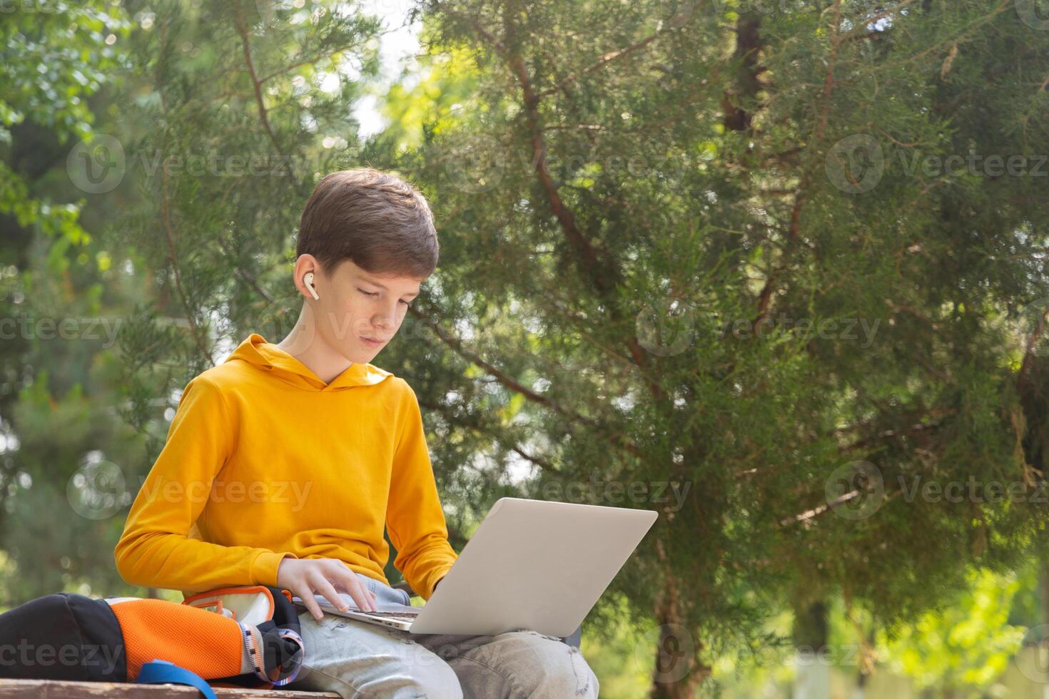 A teenage boy in a yellow hoodie sits in a park with a laptop, wearing wireless headphones. Green trees in the background create a natural summer setting. Side view with copy space photo