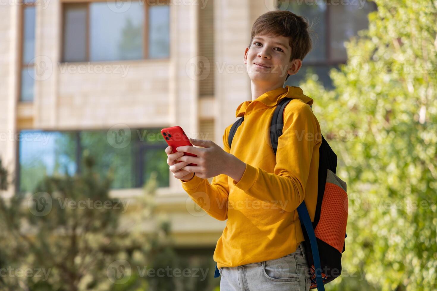 A teenage boy in a yellow hoodie holds a smartphone and looks directly at the camera. Neutral background, casual pose, and modern technology photo
