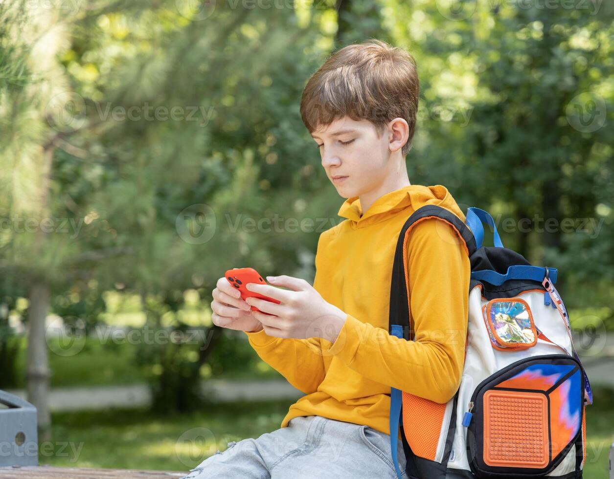 A teenage boy in a yellow hoodie sits in a park and looks at his smartphone. Green trees in the background create a relaxed outdoor setting photo