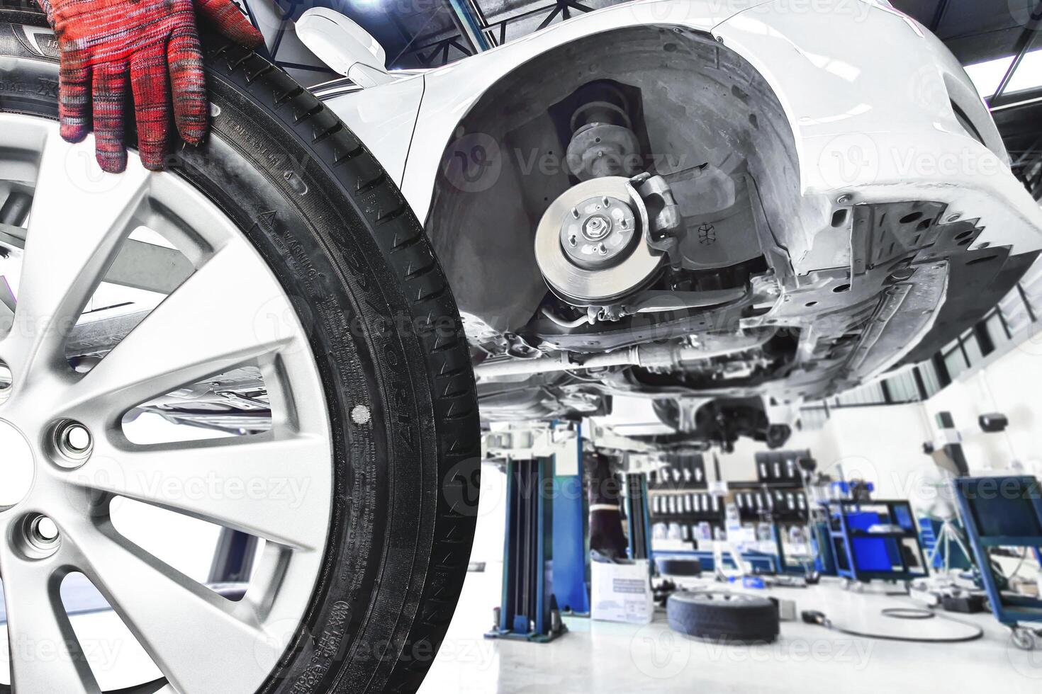 Wheel Alloy Tire Change in Auto Repair Garage, A mechanic hand holds a car wheel, removed from a white car elevated on a lift in a auto service garage. photo