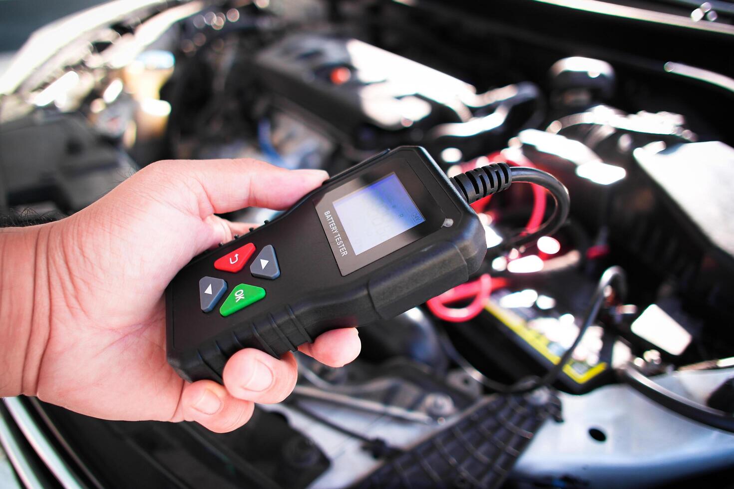 Car Battery Diagnostic Tool, A technician holds a car battery tester over an open car hood, with the engine visible, to check for vehicle battery issues. photo