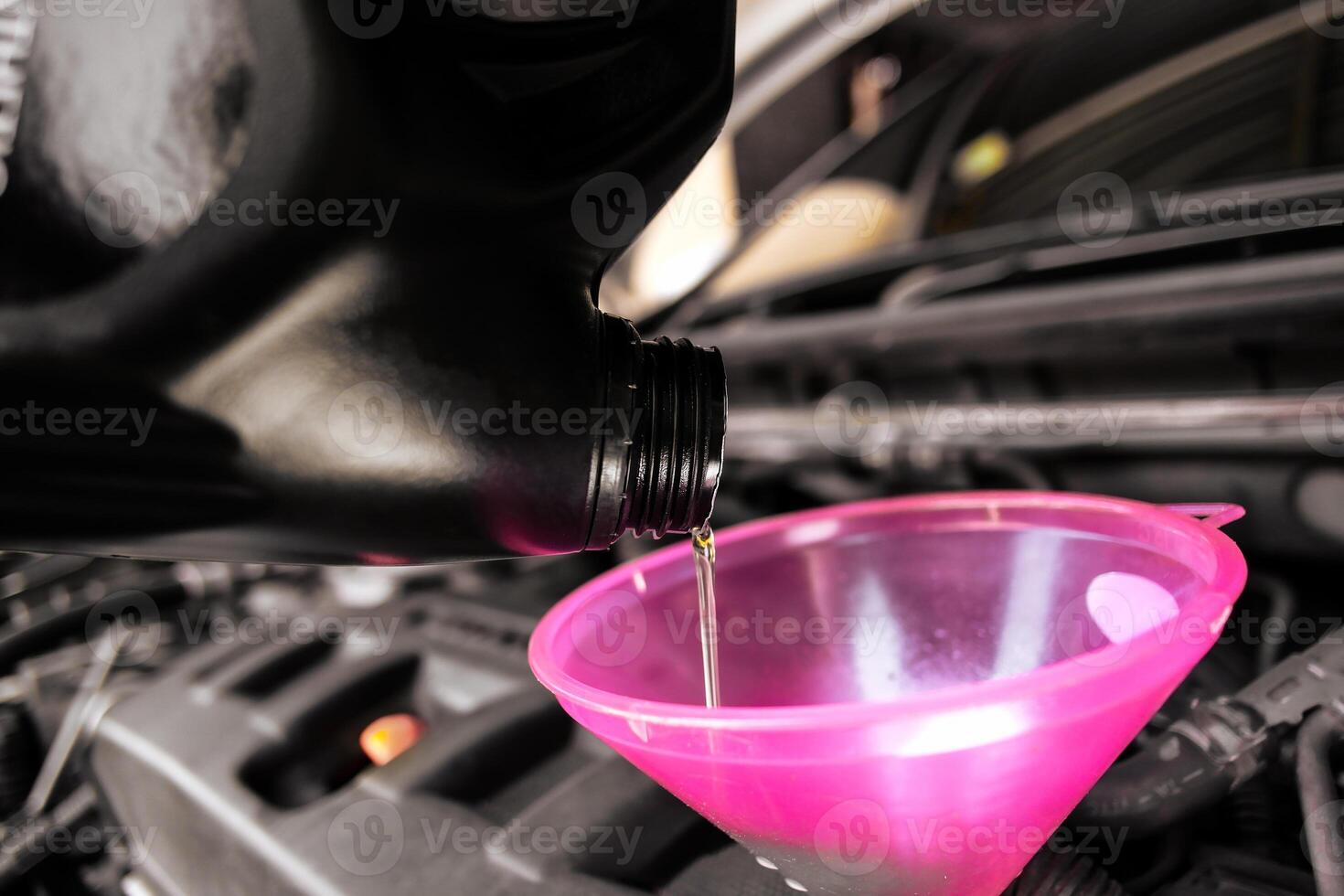 Engine oil or motor oil, is being poured from a dark bottle into a pink funnel in a car engine bay. photo