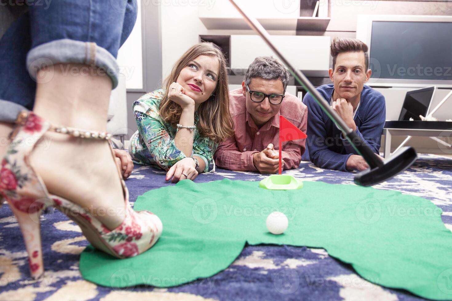 close up of a female foot in high heel shoe and a golf club with three young friends lying on the floor at home in the background photo