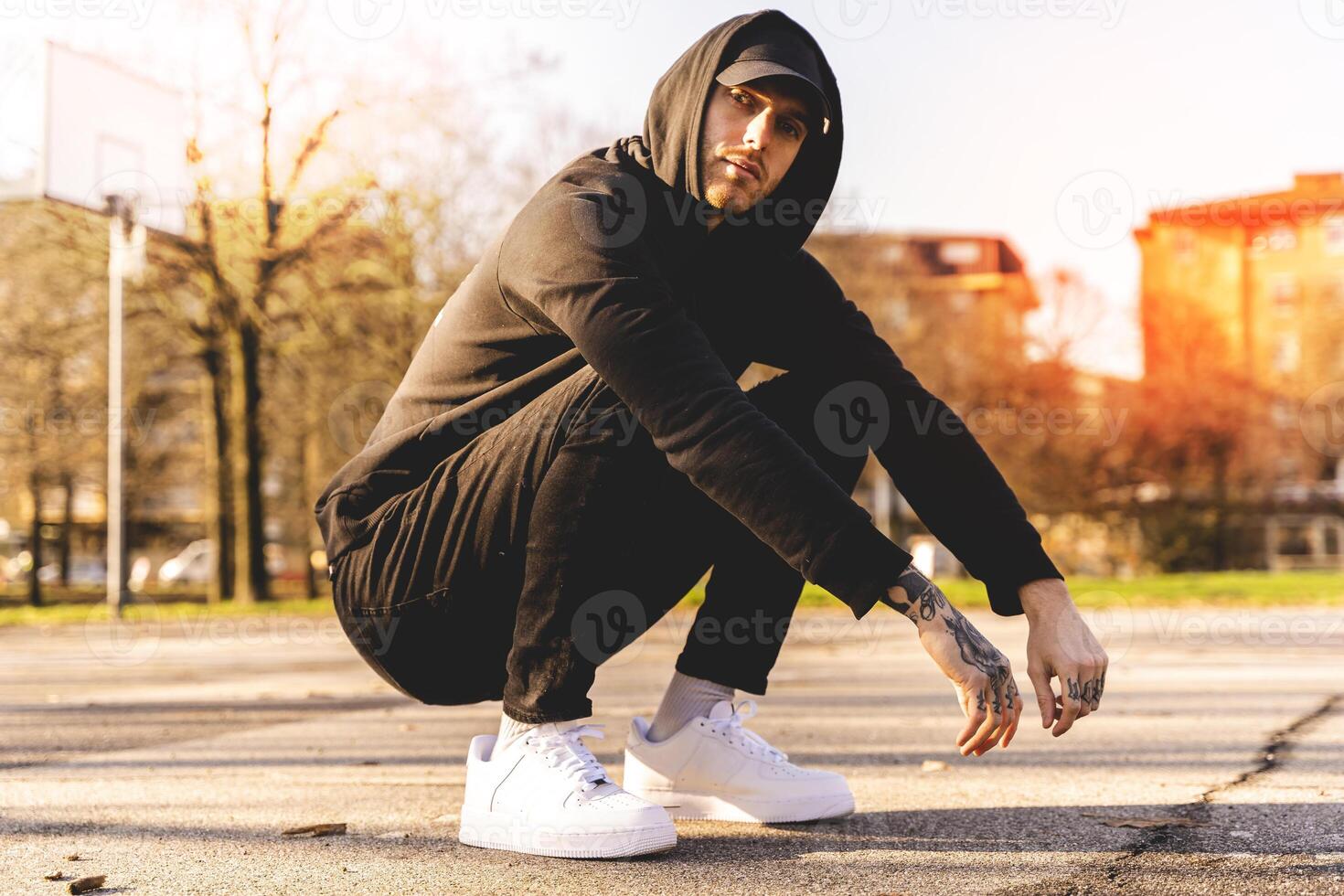 young rapper in black sweatshirt and hoodie posing in the middle of an outdoor basketball court photo
