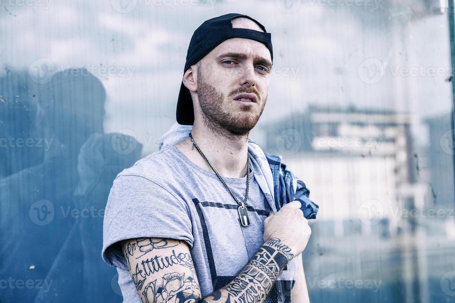 portrait of young tattooed rapper looking into camera in front of large window in the outskirts photo