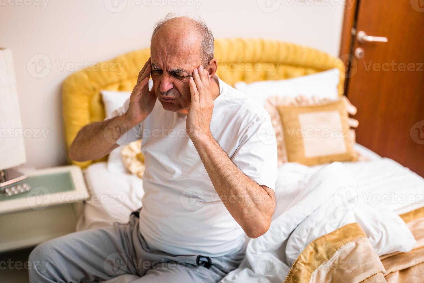 Senior man sitting on bed at dawn, holding his head in pain, struggling with a headache and exhaustion. photo