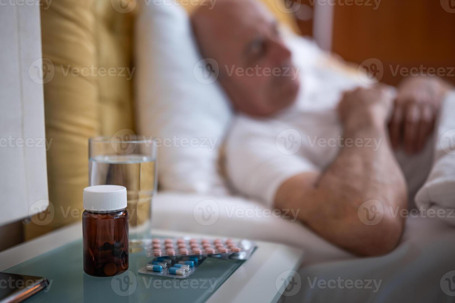Aged male peacefully sleeping in bed with pills and glass of water next to him on night table in his bedroom. Selective focus on bottle of pills. photo