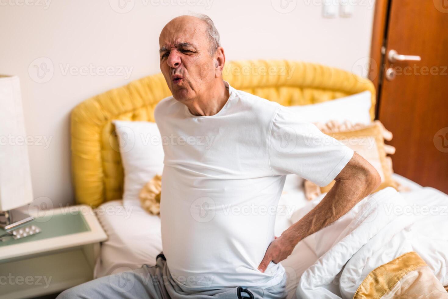 Caucasian senior male with a gray hair having pain in back after waking up and sitting on bed in the morning, representing physical injury or muscle strain in older age. photo