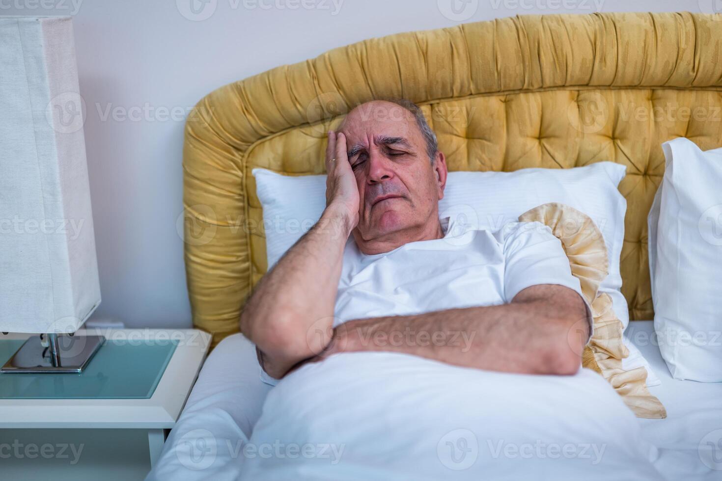 Elderly man lying in bed clutching his head in pain, suffering from a morning headache and anxiety in a dimly lit bedroom. photo