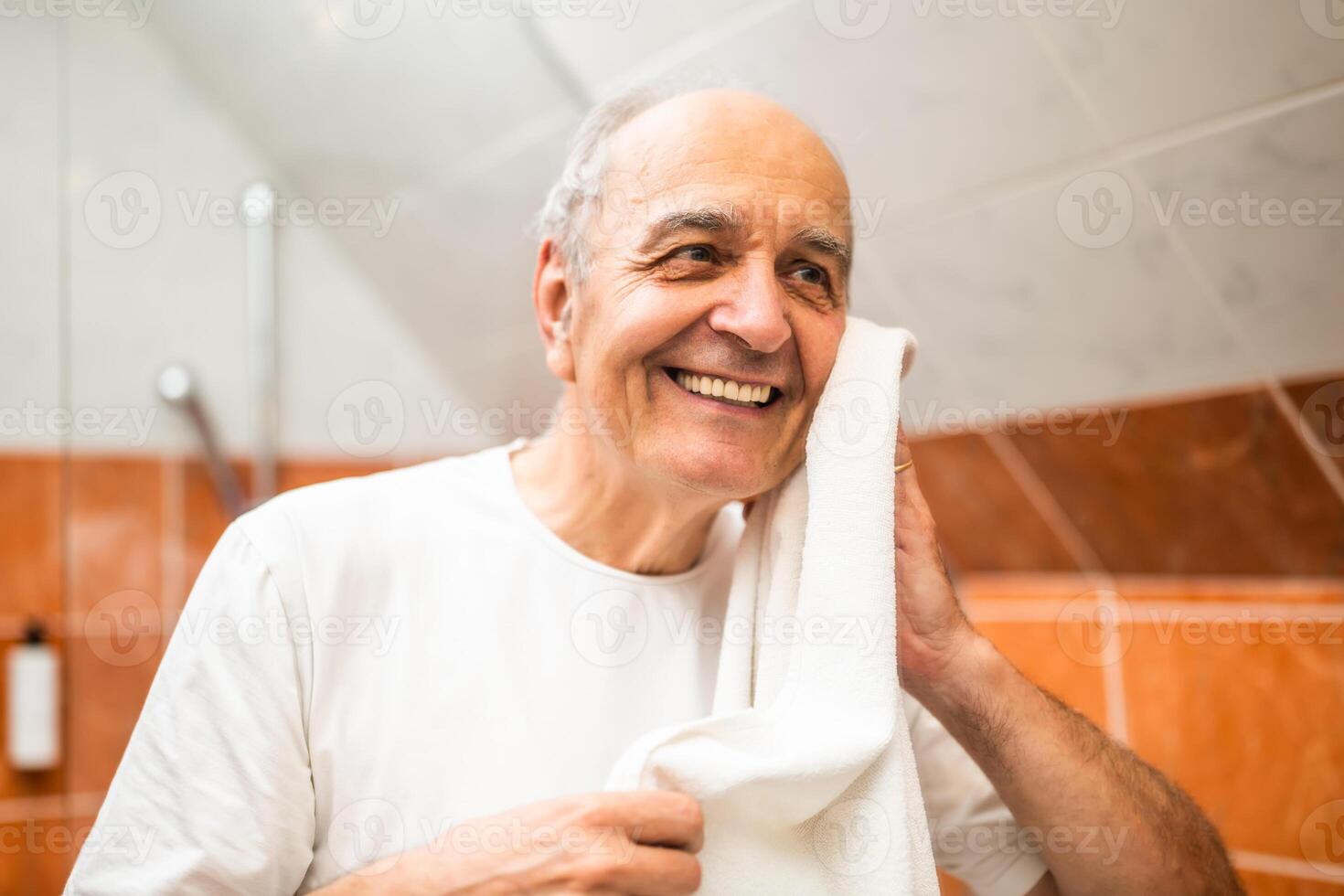 Elderly man with gray hair smiling as he dries his face with a towel in front of the bathroom mirror during his morning skincare routine. photo