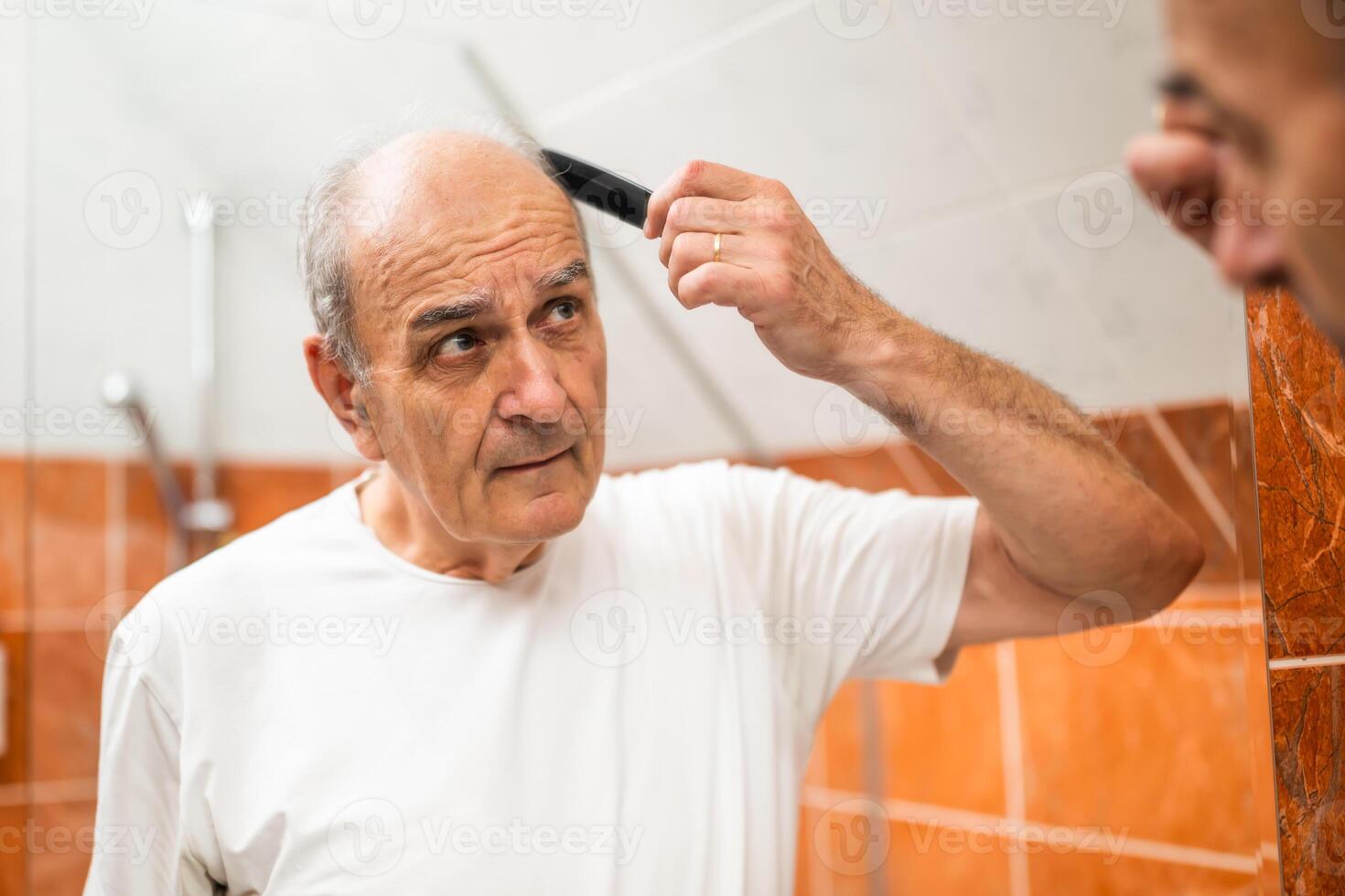 Elderly man combing his sparse gray hair while appearing distraught, highlighting the emotional toll of hair loss and aging. photo