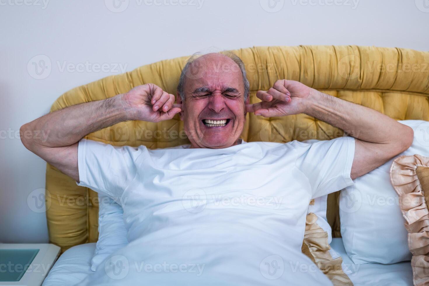 Frustrated and tired old man in pajamas lying in bed and covering ears, suffering from insomnia due to loud noise in the early morning. photo