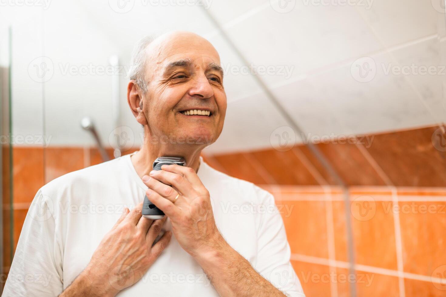 Balding mature man with a cheerful expression engaging in his daily body care by shaving his beard with electric razor in bathroom. photo
