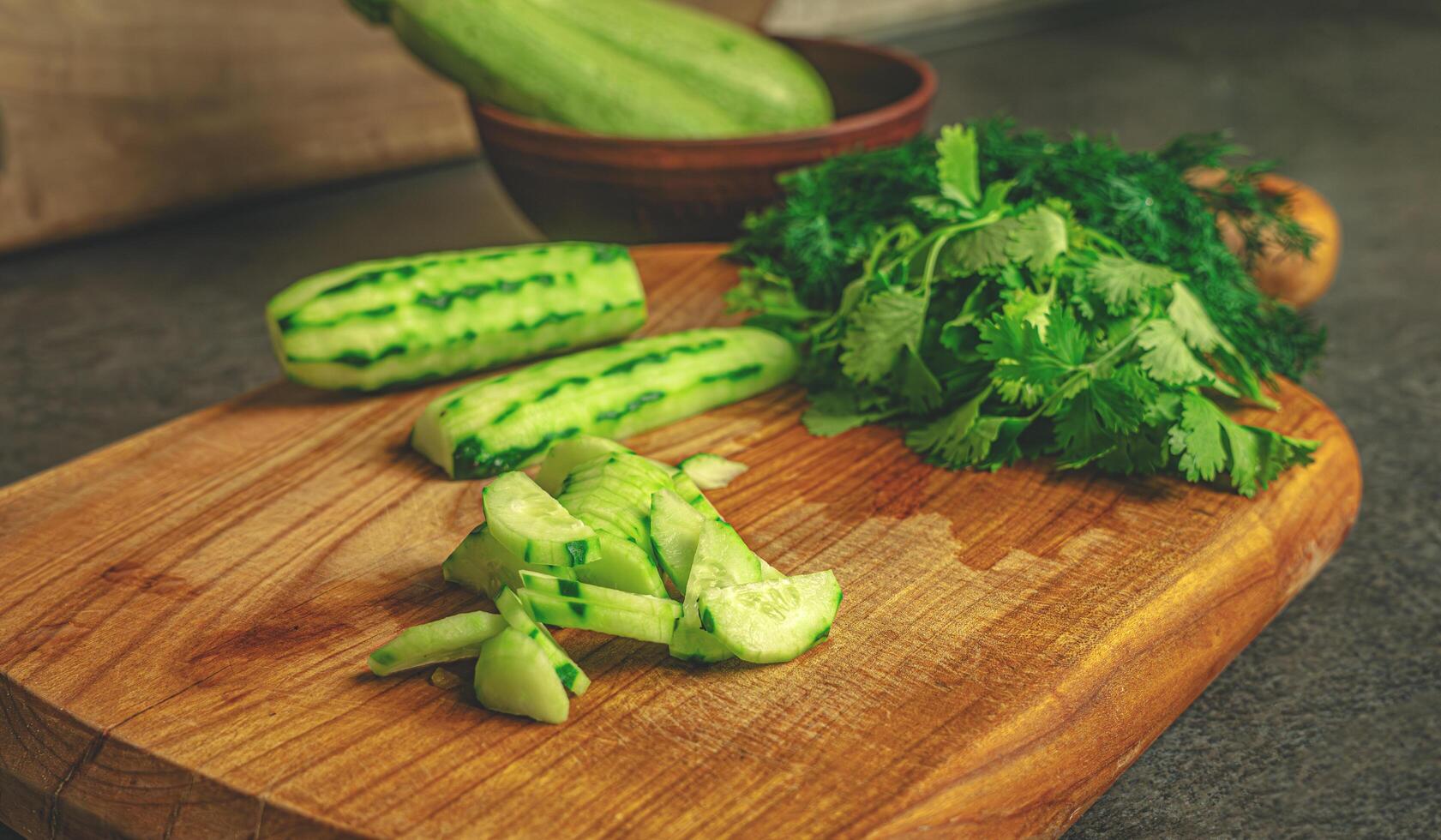 Sliced cucumbers and aromatic greens are arranged on a warm-toned wooden cutting board made of cherry wood. The scene evokes a sense of simplicity, freshness, and natural flavor photo