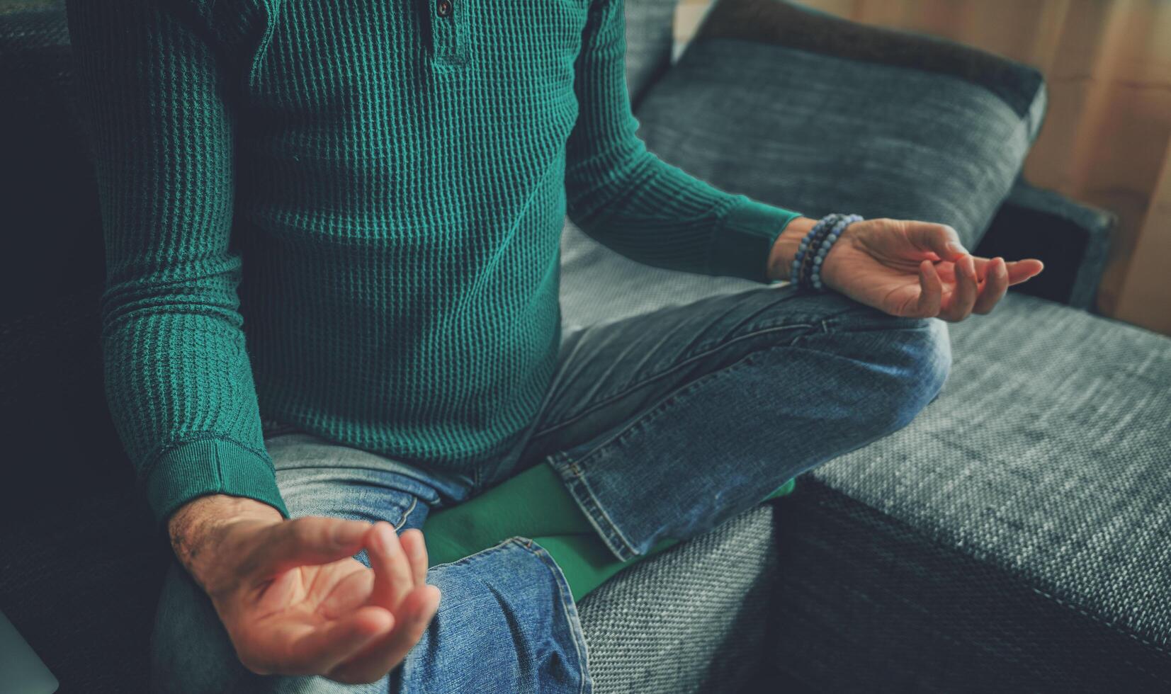 A middle-aged man sits peacefully in lotus pose on a sofa, wearing a green long-sleeve sweater and jeans. Natural stone bracelets accent his relaxed, mindful appearance. The home setting conveys photo