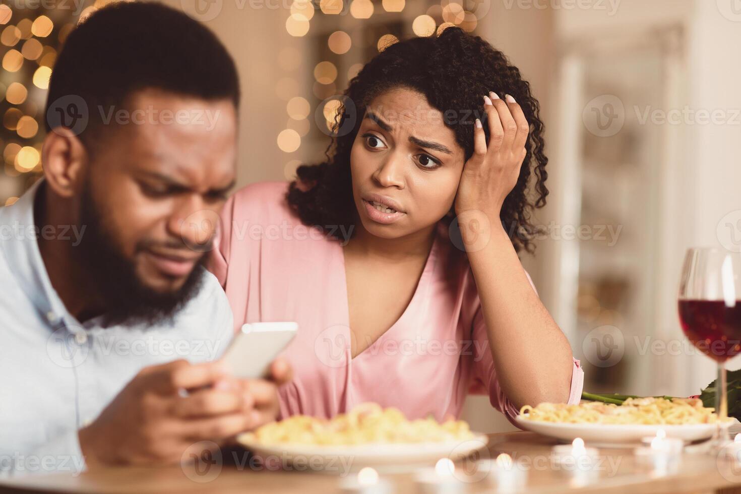 Phubbing. Angry young black woman looking at her boyfriend stuck in smartphone, unhappy about neglect and lack of communication. Jealous lady and gadget addicted man having dinner in cafe or at home photo