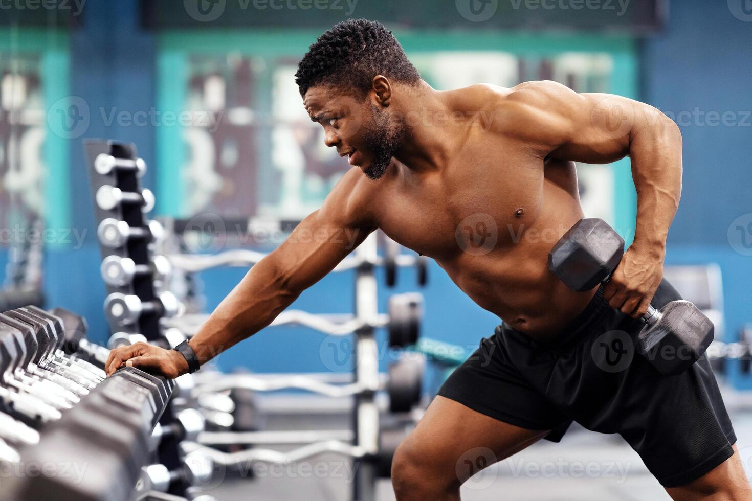 Motivated african american shirtless bodybuilder training arms and back muscles with dumbbells at modern gym, empty space. Hot black young man working on his biceps, lifting barbell with one hand photo