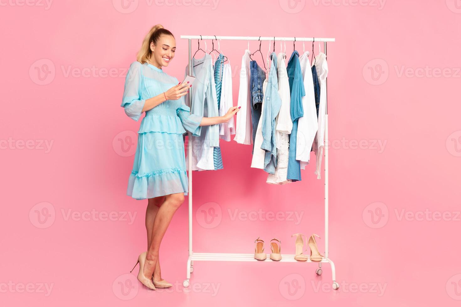 Fashion Concept. Full length vertical portrait of smiling female stylist holding and using smartphone, texting sms or making purchases in online store standing near rack with clothes at pink studio photo