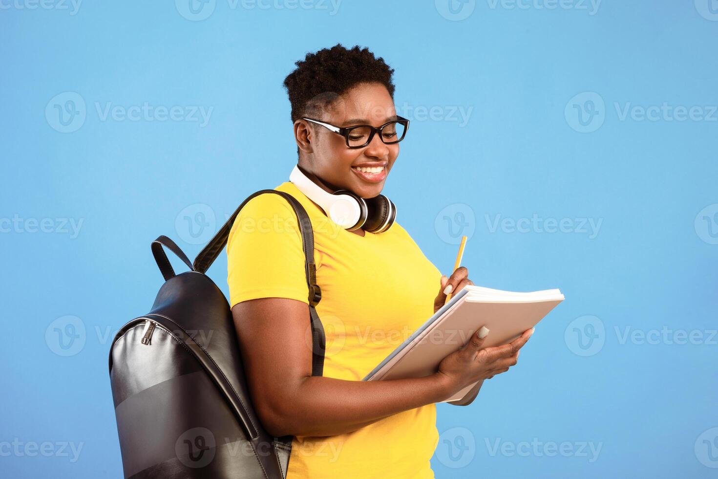 Black Plus-Size Student Lady Learning Posing Holding Books Standing Over Blue Studio Background, Wearing Eyeglasses And Taking Notes. Knowledge, College And University Study Concept photo