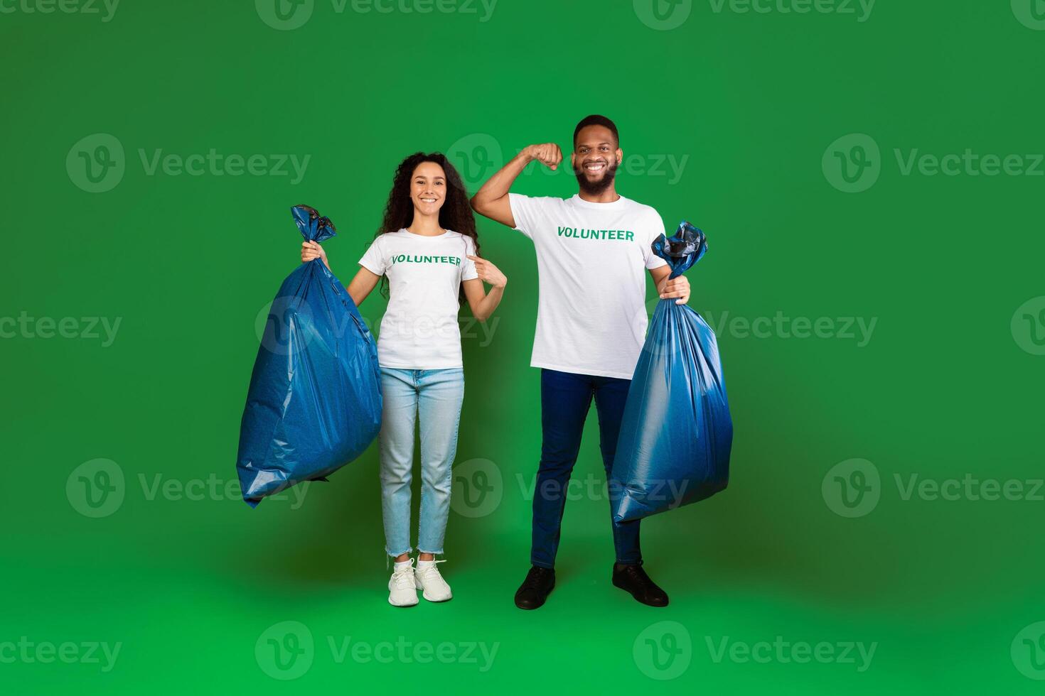Two Diverse Volunteers Holding Blue Garbage Bags Showing Biceps And Pointing At Volunteer Inscription On T-Shirts Posing Over Green Studio Background. Keep Environment Clean. Full Length photo