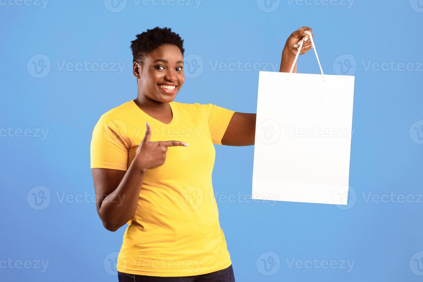 Great Sales. Happy Black Woman Showing Shopping Bag Pointing Finger At Blank White Shopper Handbag Standing Over Blue Studio Background, Smiling To Camera. Discounts Offer Advertisement. Mockup photo