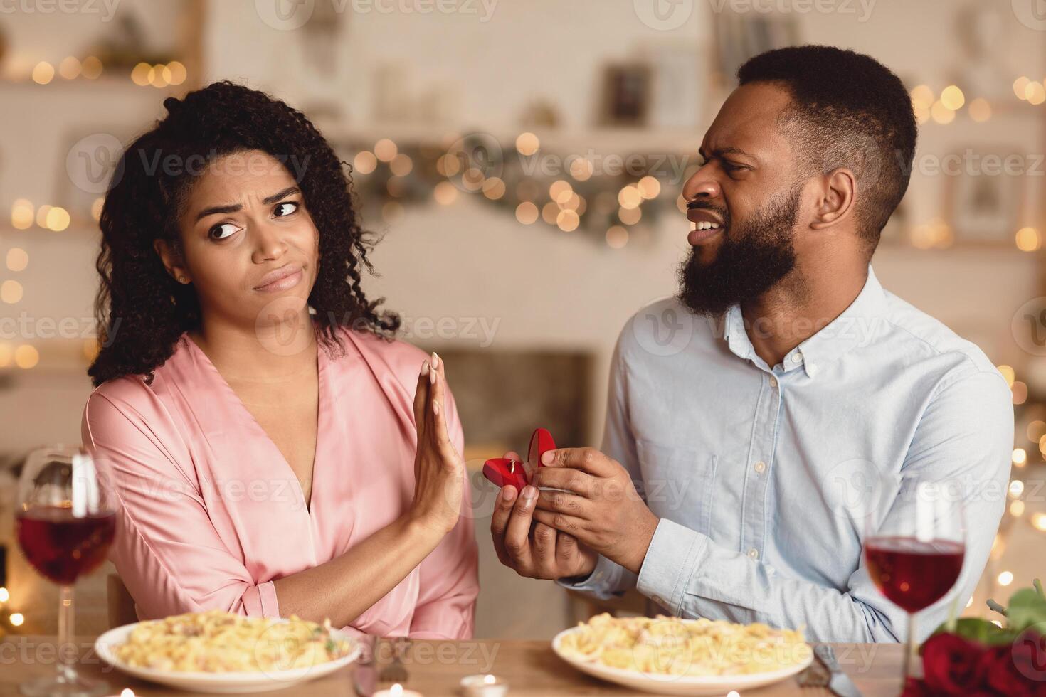 Proposal Fail. Portrait of unhappy african american man proposing to his gilfriend, holding open box with marriage ring, uninterested bored woman saying no, showing stop gesture with palm photo