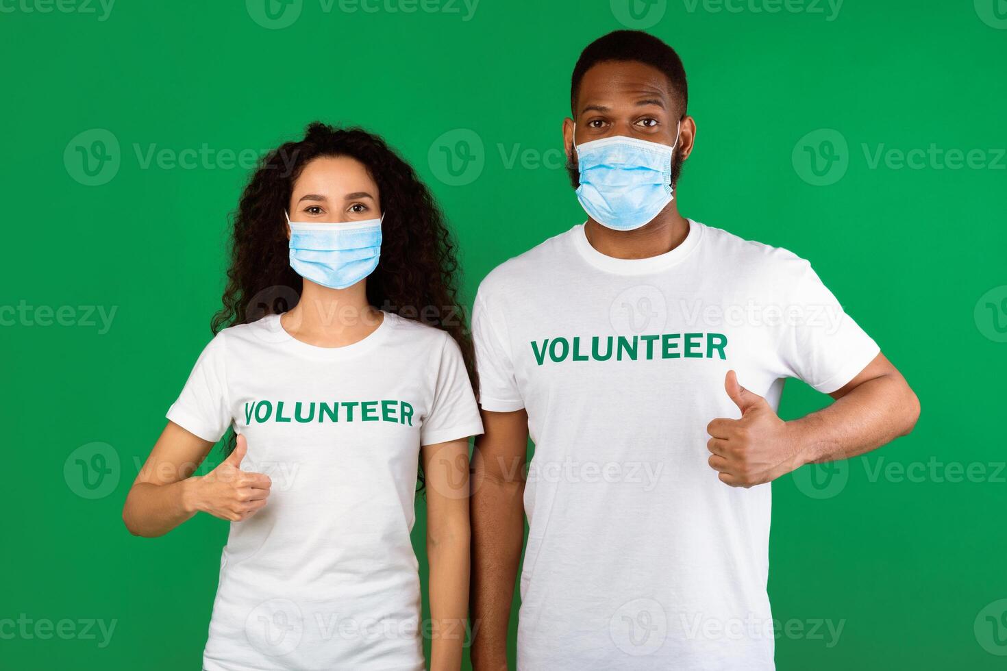 Couple Of Volunteers Pointing Fingers At Face Masks And Gesturing Thumbs Up Standing Over Green Studio Background. Coronavirus Covid-19 Protection, Volunteering Work During Pandemic photo