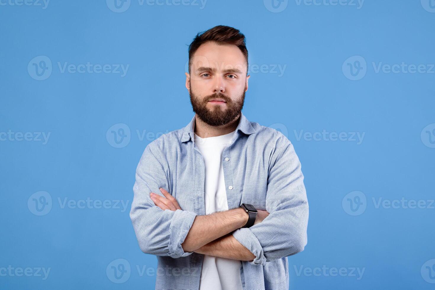 Portrait of serious millennial guy posing with crossed arms and looking at camera on blue studio background. Confident bearded young man in casual clothes having cool face expression photo