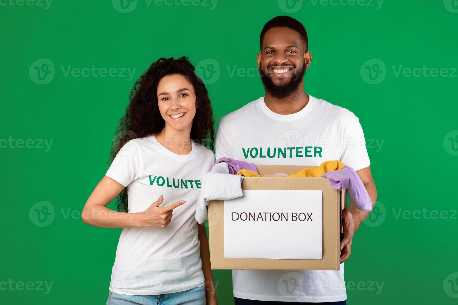 Two Multiracial Volunteers Holding Donation Container Box For Charity Standing Over Green Background In Studio. Donate Advertisement Banner. Volunteering, Voluntary Work Concept photo
