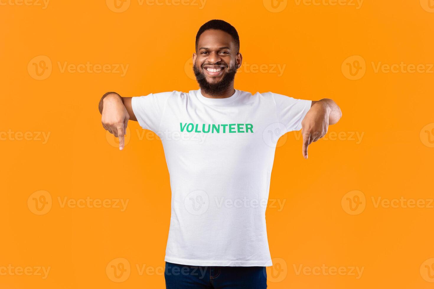 African Volunteer Guy Pointing Fingers Down Posing Smiling To Camera Over Yellow Background. Activist Advertising Something Standing In Studio. Volunteering Advertisement. Look There Concept photo