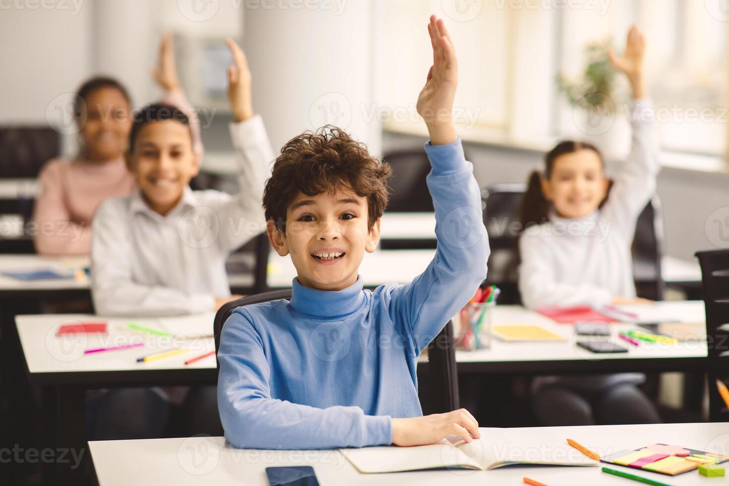 Education, Junior School, Learning and People Concept. Happy schoolboy raising hand for an answer, sitting at desk in classroom with diverse group of smiling classmates, studying with pleasure photo