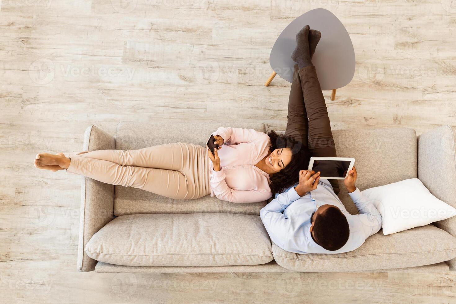 Above Top Overhead High Angle View Of African American Couple Sitting On Couch, Woman Using Smartphone, Man Holding Digital Tablet Browsing Internet Together At Home. Modern Lifestyle Concept photo