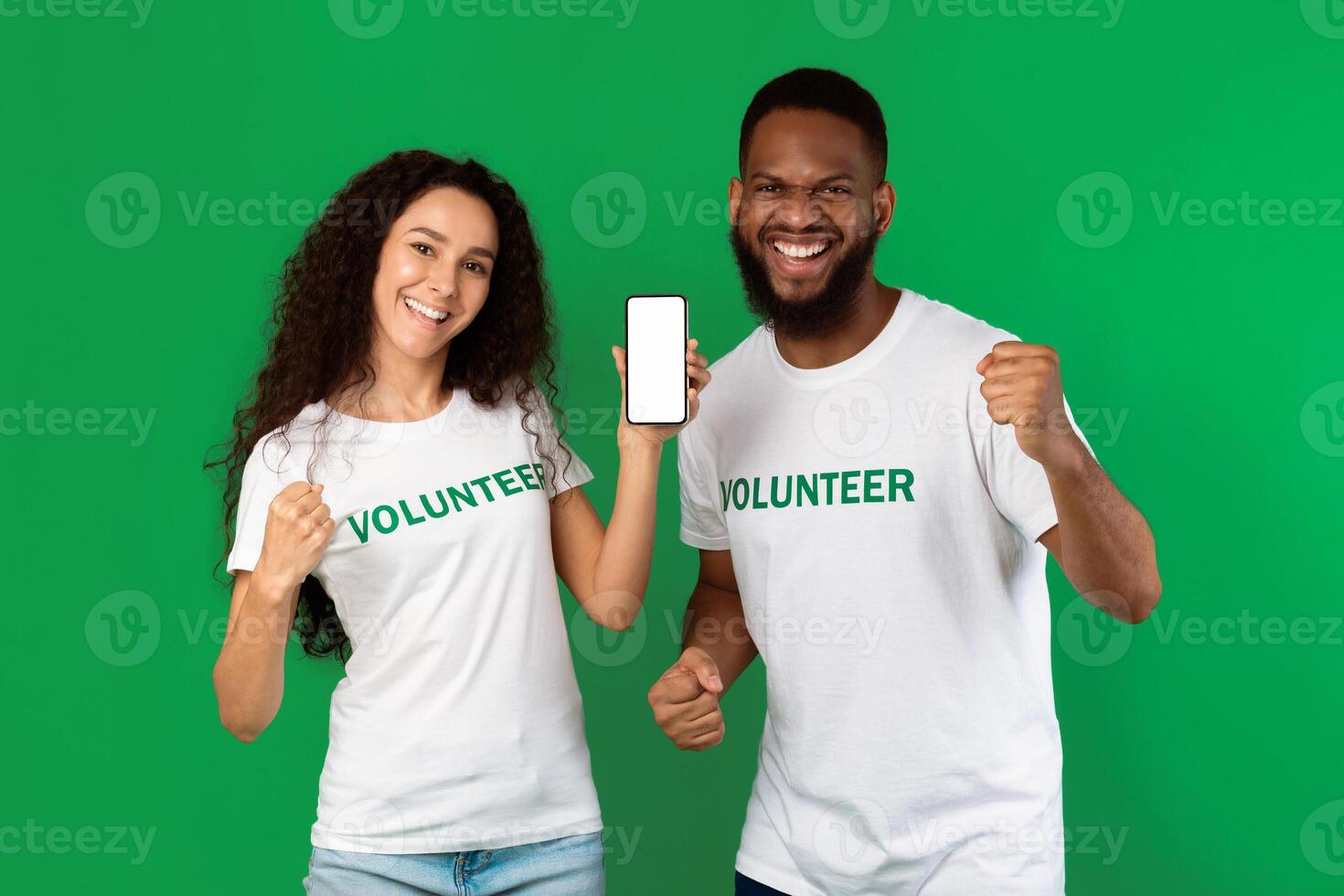 Two Mixed Volunteers Showing Mobile Phone Blank Screen Gesturing Yes In Joy Recommending Volunteering Application Standing On Green Background. Studio Shot. Call Us Concept. Mockup photo