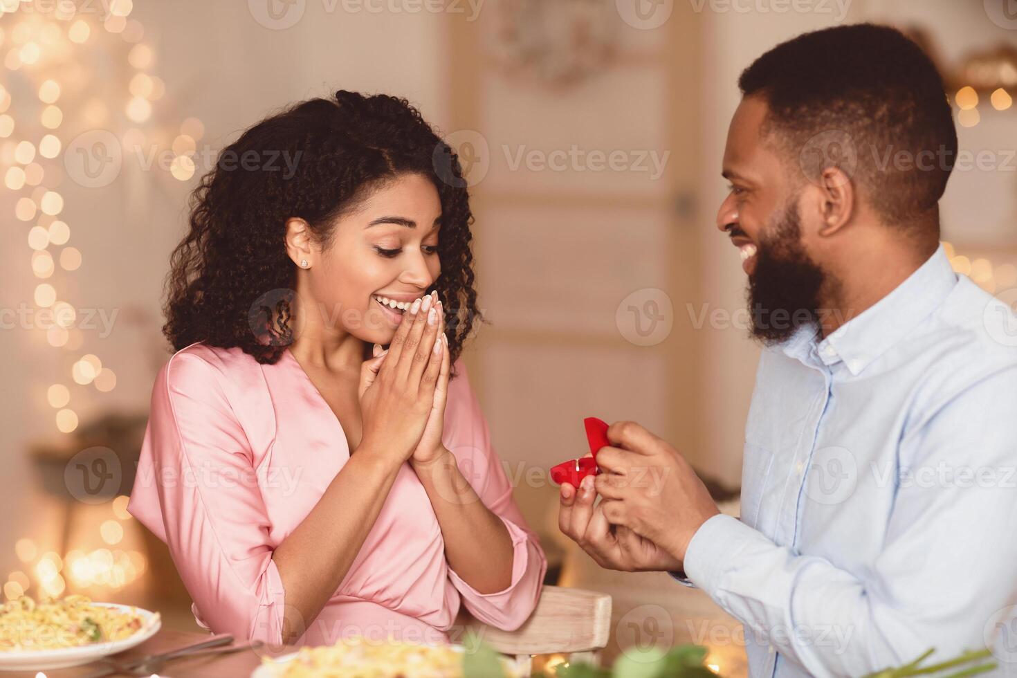Marry Me. Smiling bearded african american guy holding and showing open box, offering marriage ring to his surprised excited fiance, getting engaged in restaurant or at home at romantic dinner photo