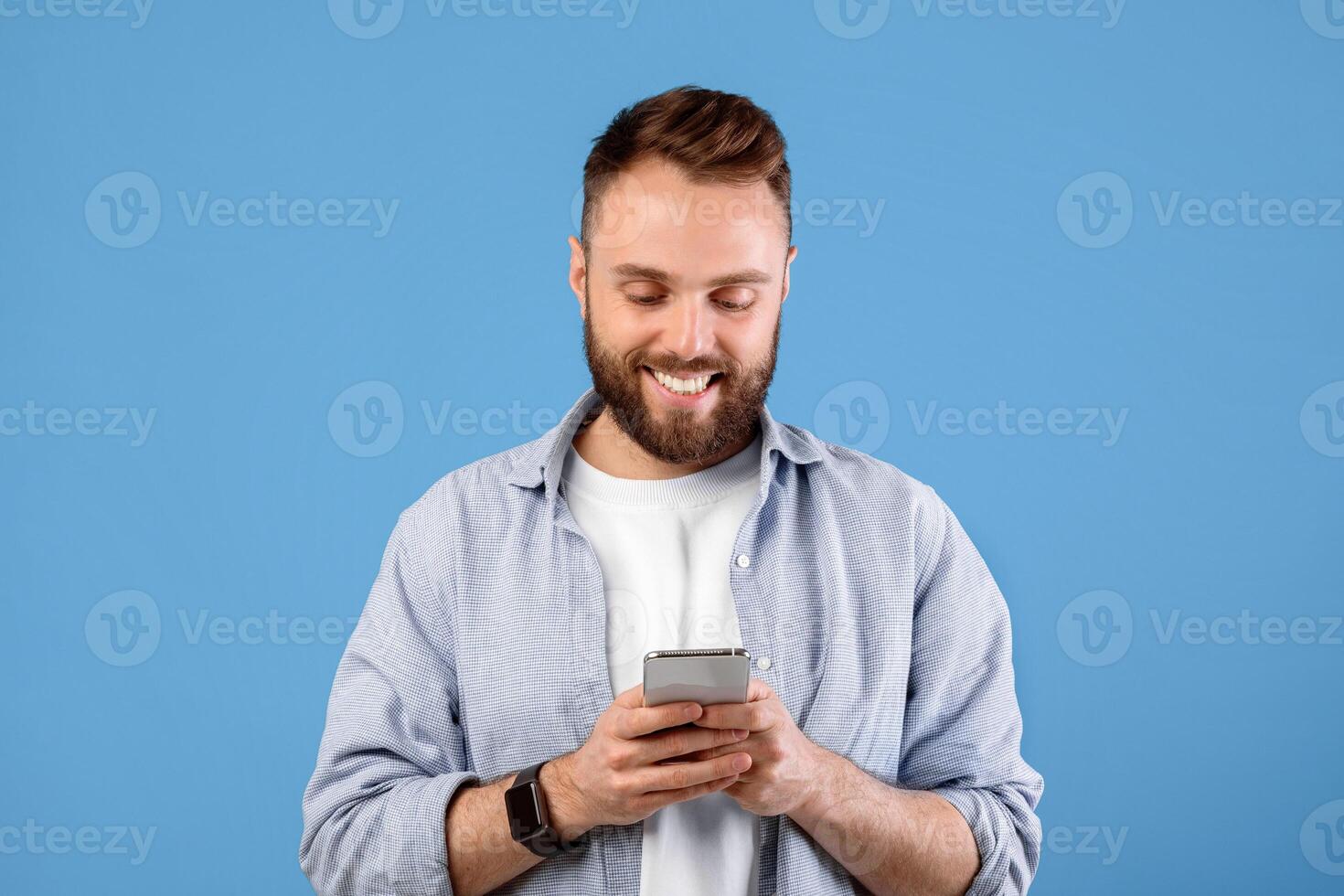 Online life soncept. Funky bearded man using cellphone on blue studio background. Positive young guy with modern gadget surfing internet, watching videos, posting in social media photo
