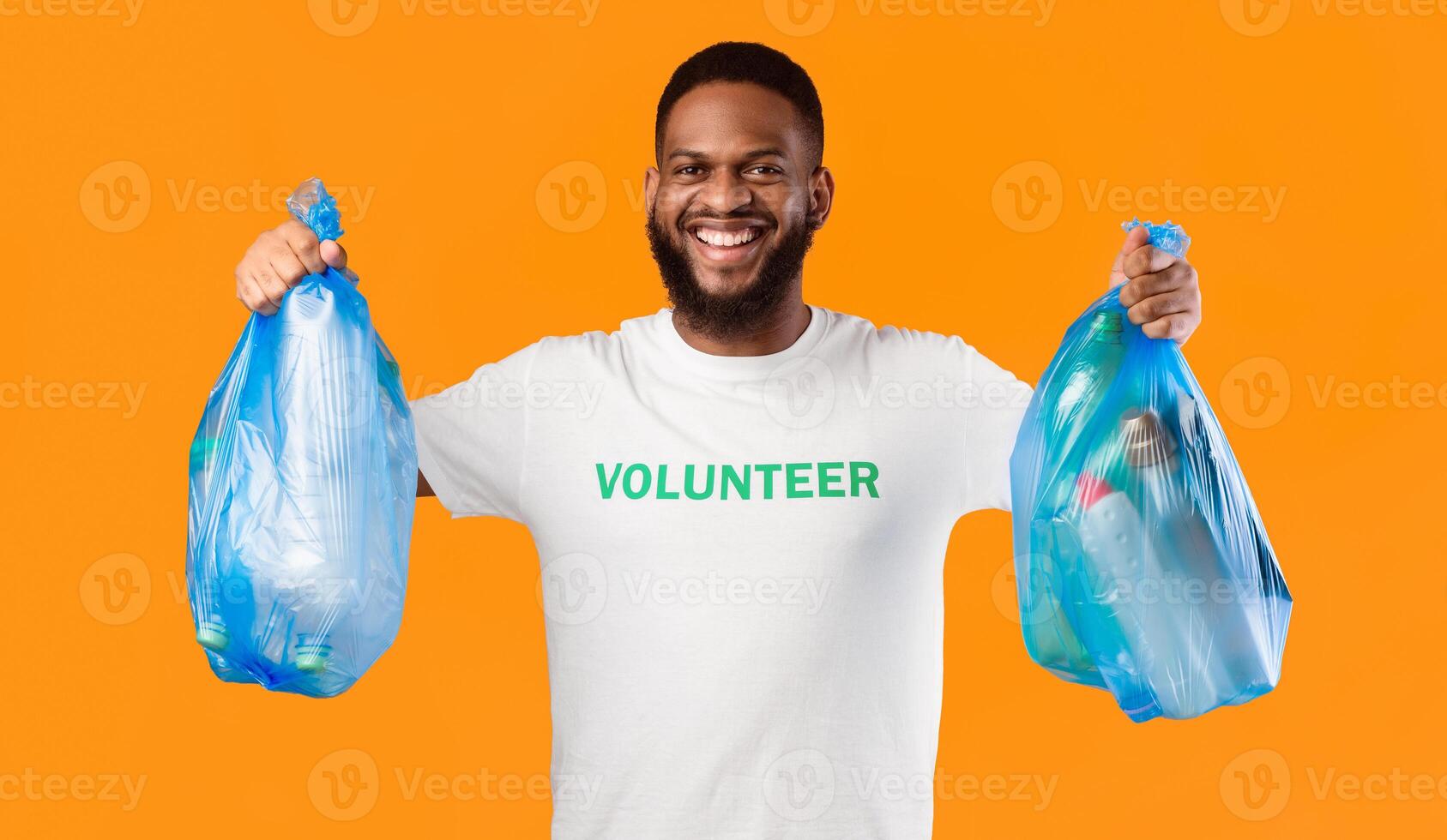 Black Volunteer Guy Holding Garbage Bags With Used Plastic Litter And Bottles Posing Smiling To Camera Standing On Yellow Background. Clean Environment, Recycling And Waste Sorting, Volunteering Work photo