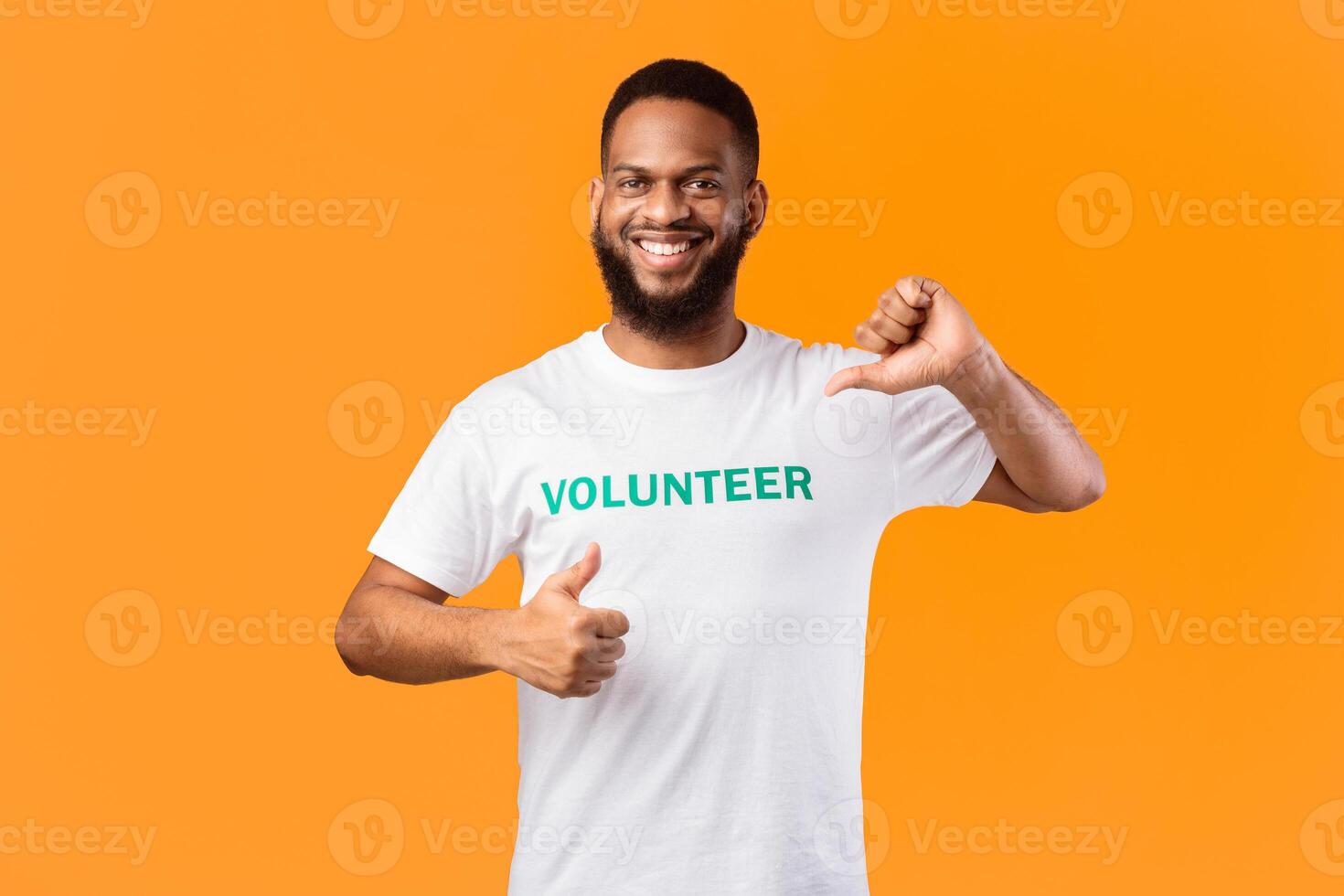 I like volunteering. Smiling african volunteer man pointing thumb at himself gesturing thumbs-up posing on yellow studio background, smiling to camera. Activist's portrait photo