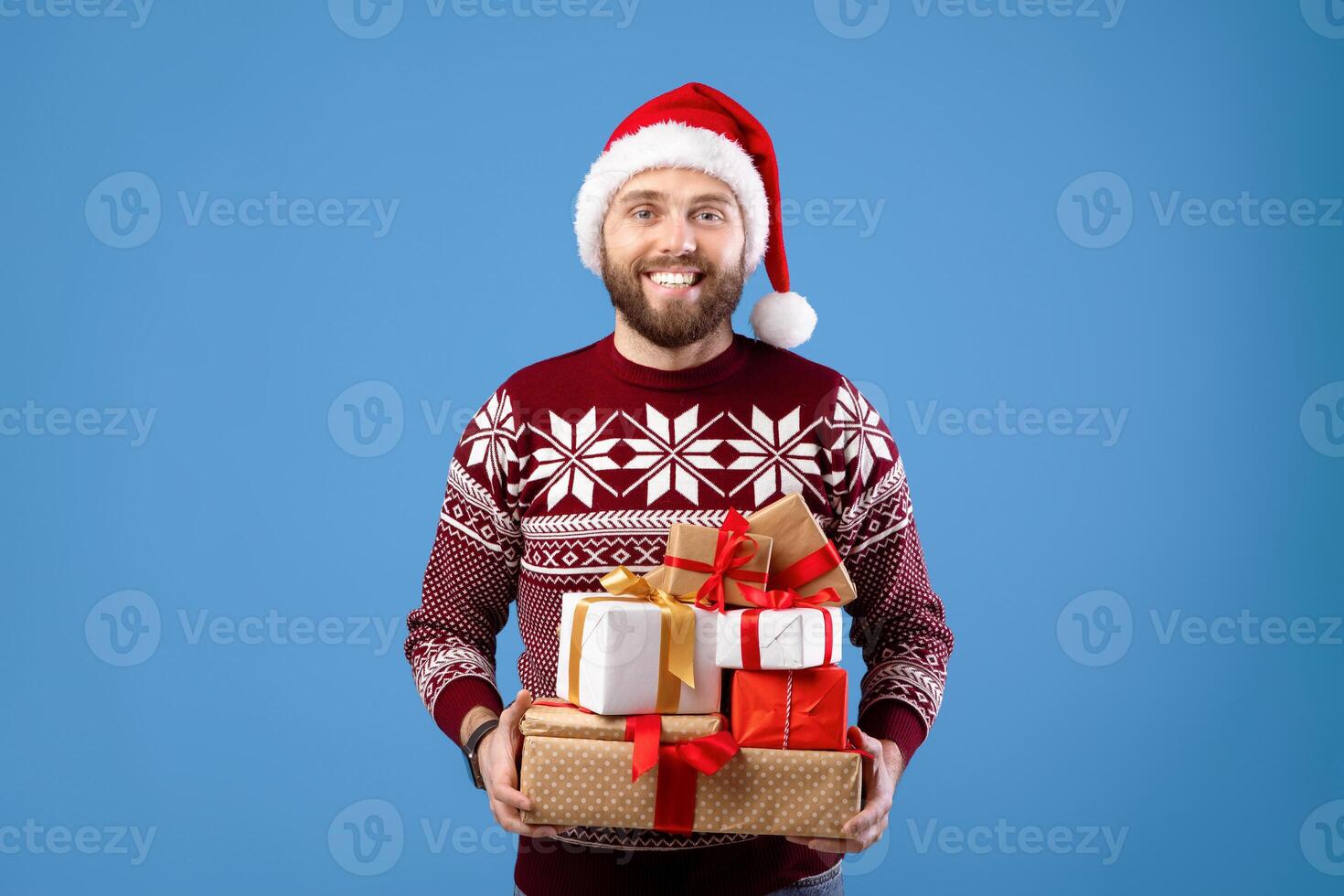 Portrait of joyful young guy in woolen sweater and Santa hat holding stack of gifts over blue studio background. Bearded millennial man with heap of presents celebrating New Year photo